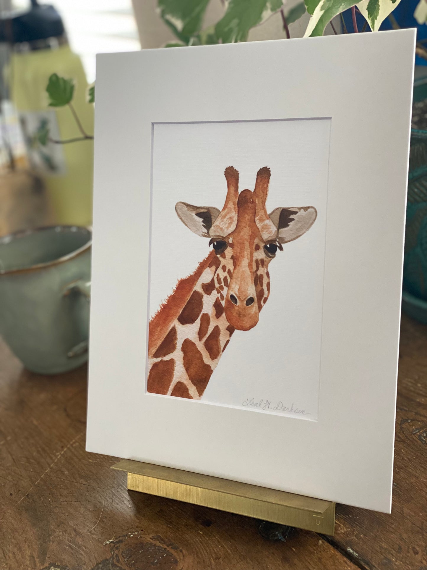 matted 5x7 print of watercolor giraffe painting sitting on a gold picture holder.