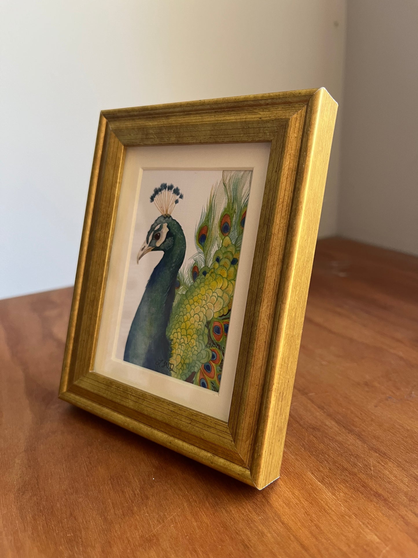 Three quarter view of a watercolor peacock in a gold picture frame.