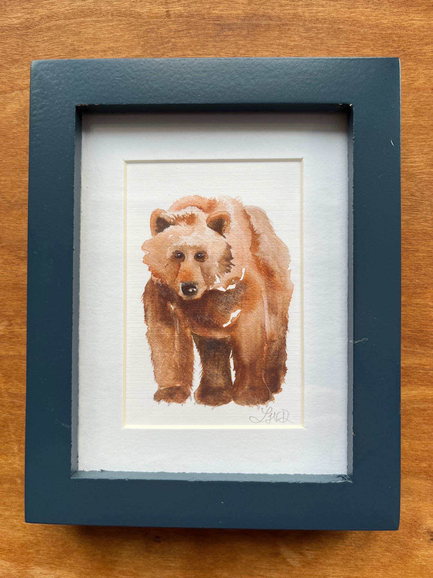 Watercolor painting of a bear in a blue picture frame with a white mat.