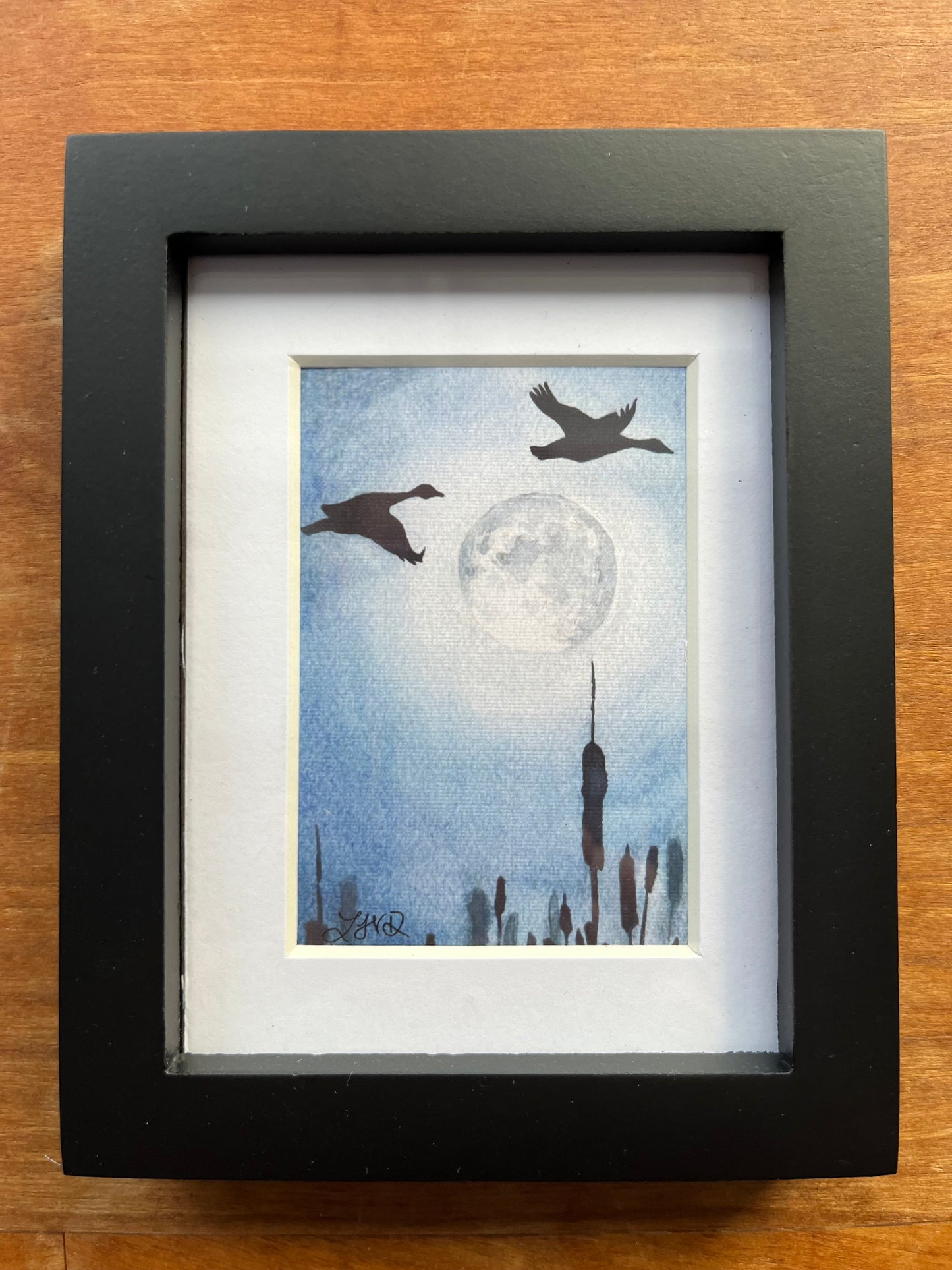 Watercolor painting of silhouetted gees flying at night by a full moon in a black picture frame.