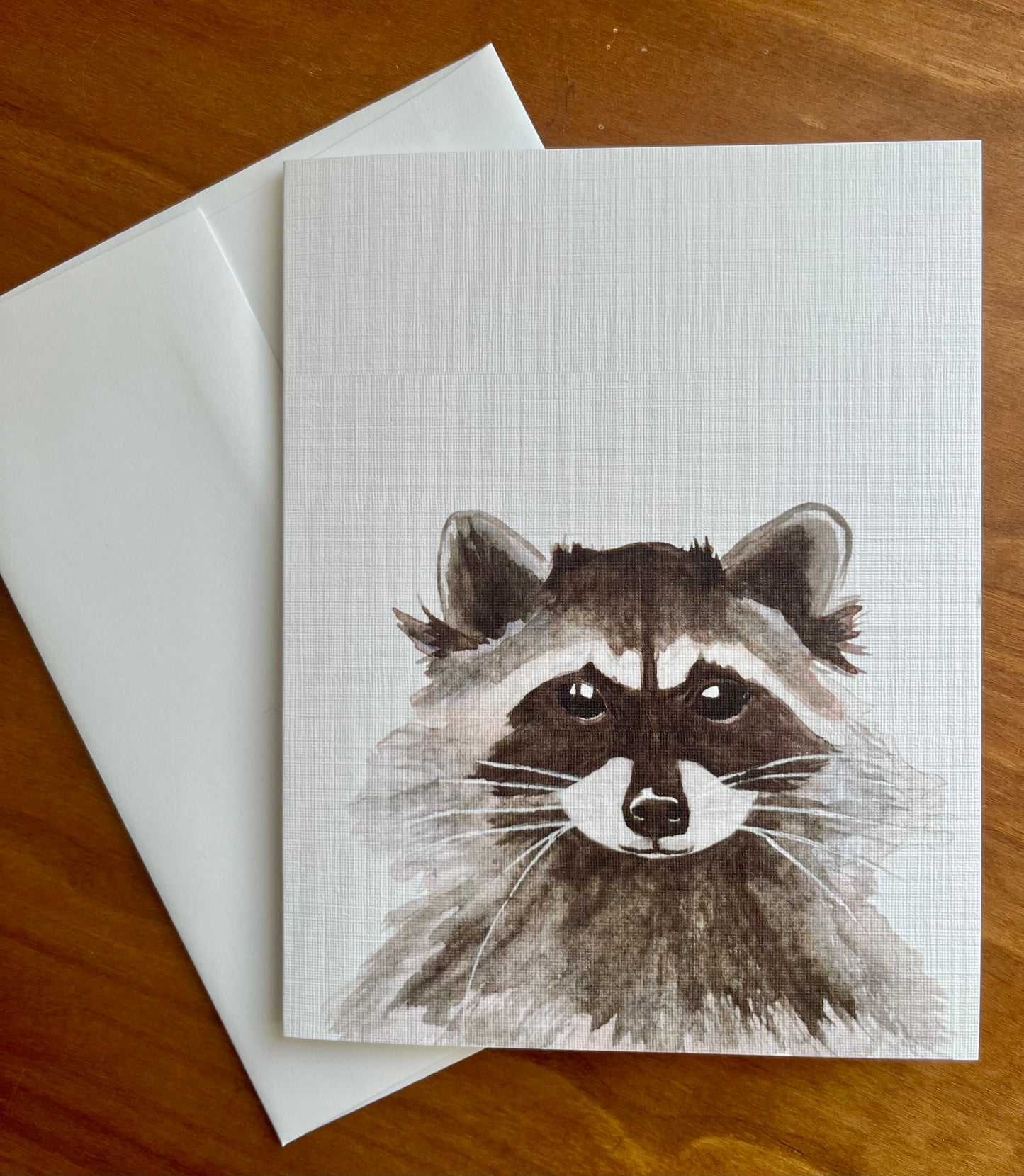 Greeting card with a watercolor painting of a raccoon along with a white envelope.