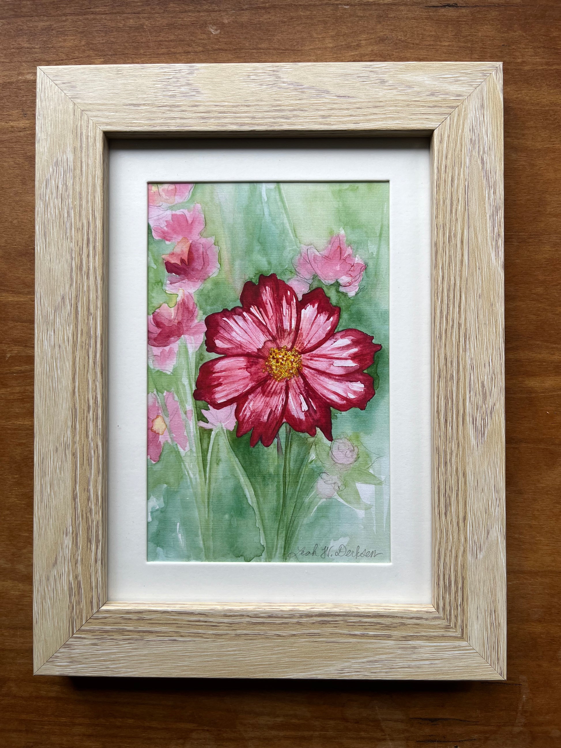 Watercolor painting of pink cosmos flowers in a blonde wood picture frame.