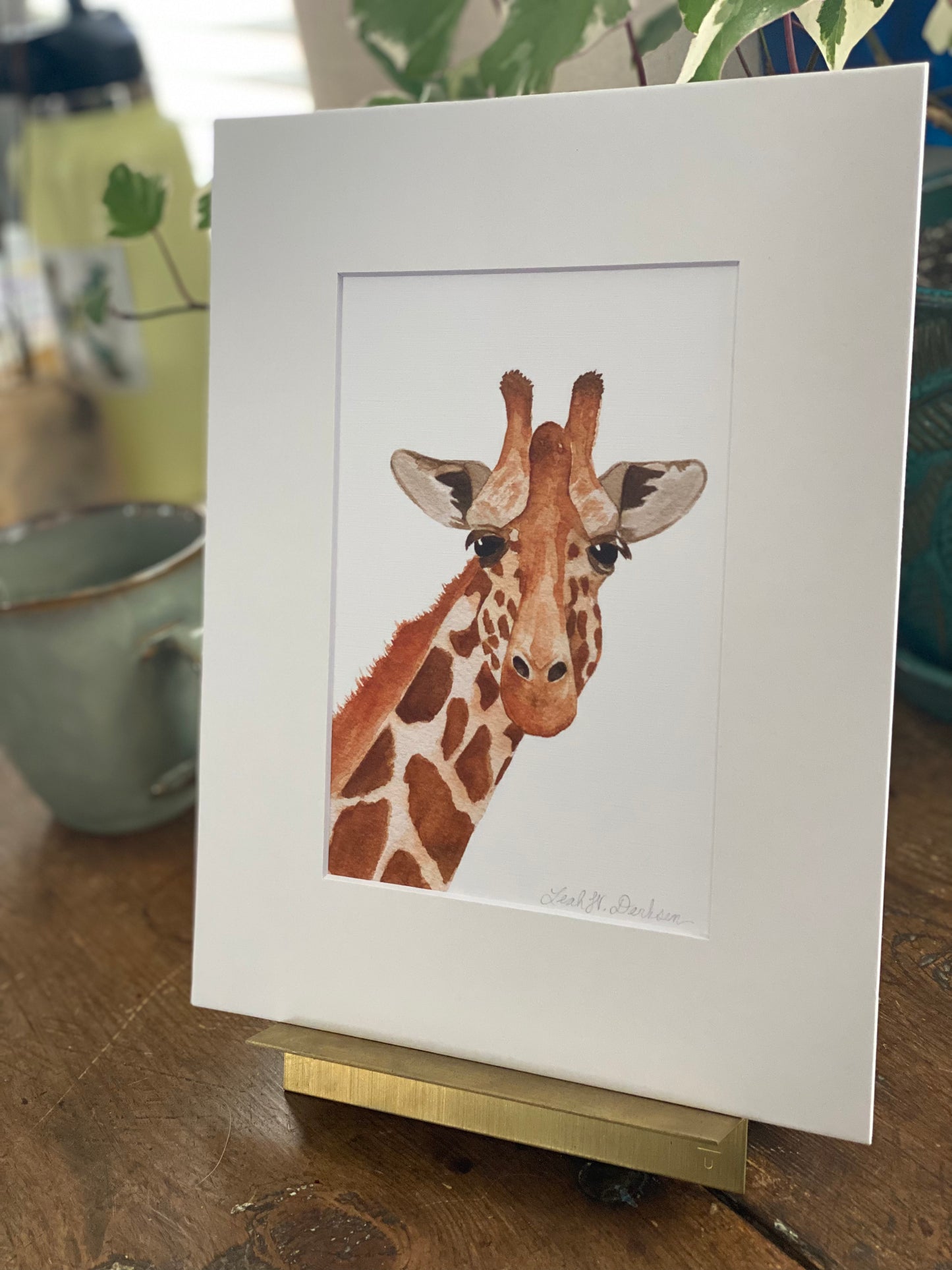 matted 5x7 print of watercolor giraffe painting sitting on a gold picture holder. 