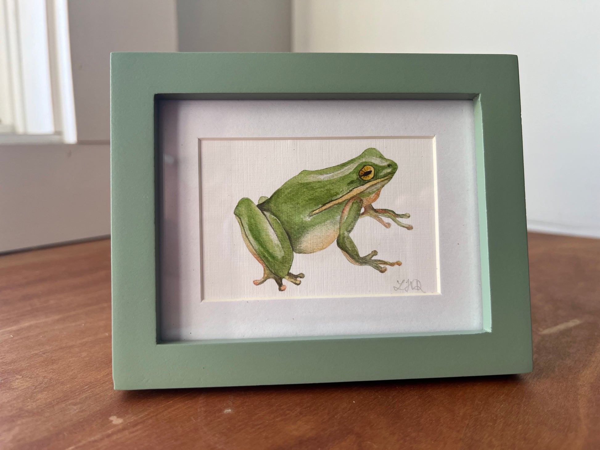 Green frog watercolor in a small green picture frame. 