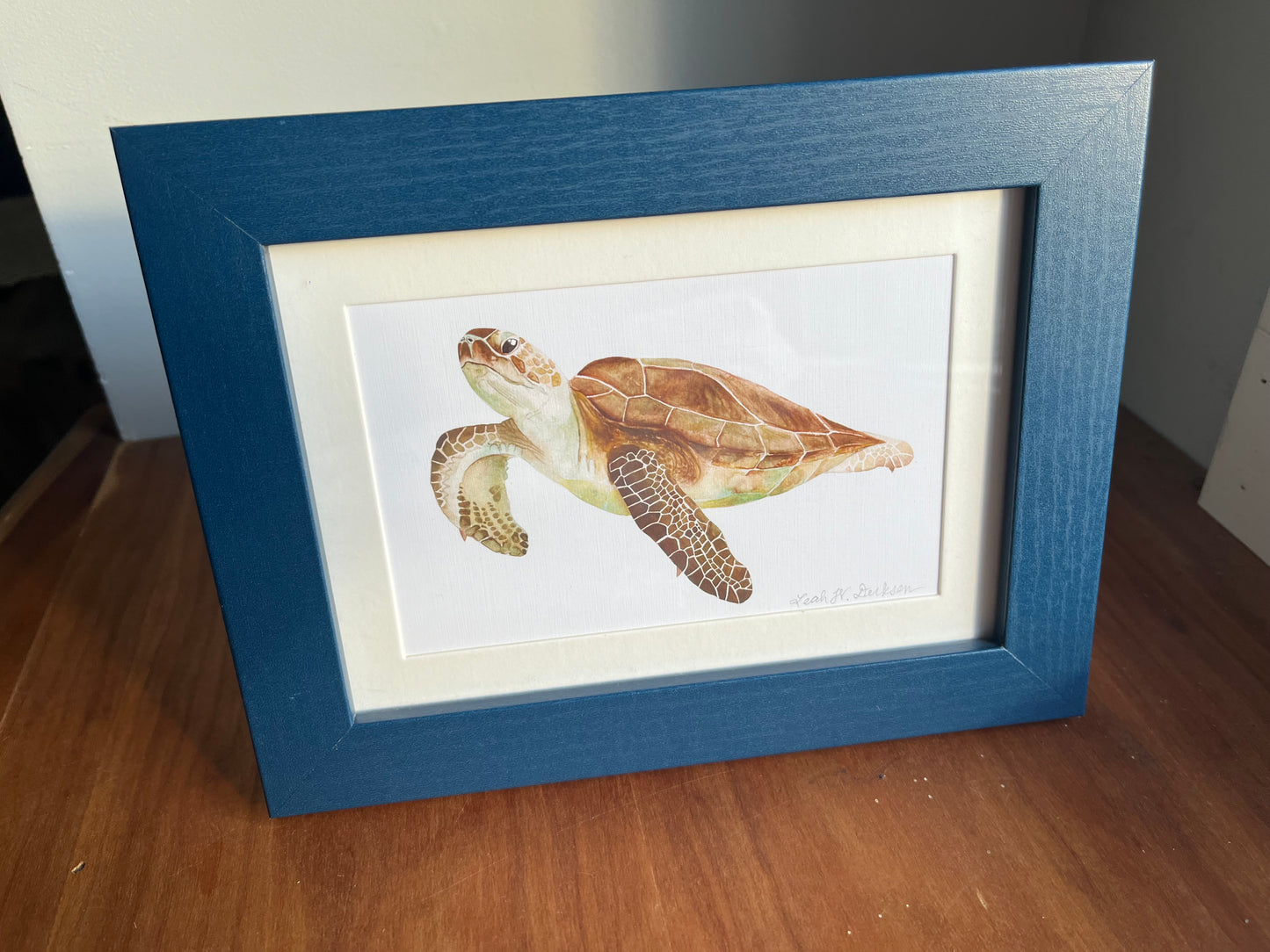 Watercolor painting of a sea turtle in a blue frame standing on a table top. 