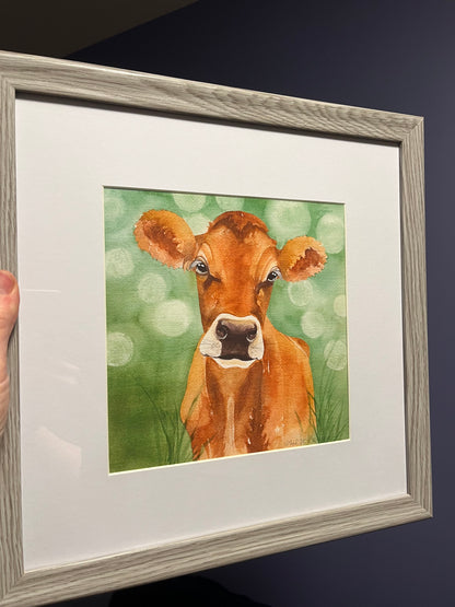 Watercolor painting of a jersey cow with a green painted background in a grey picture frame. 