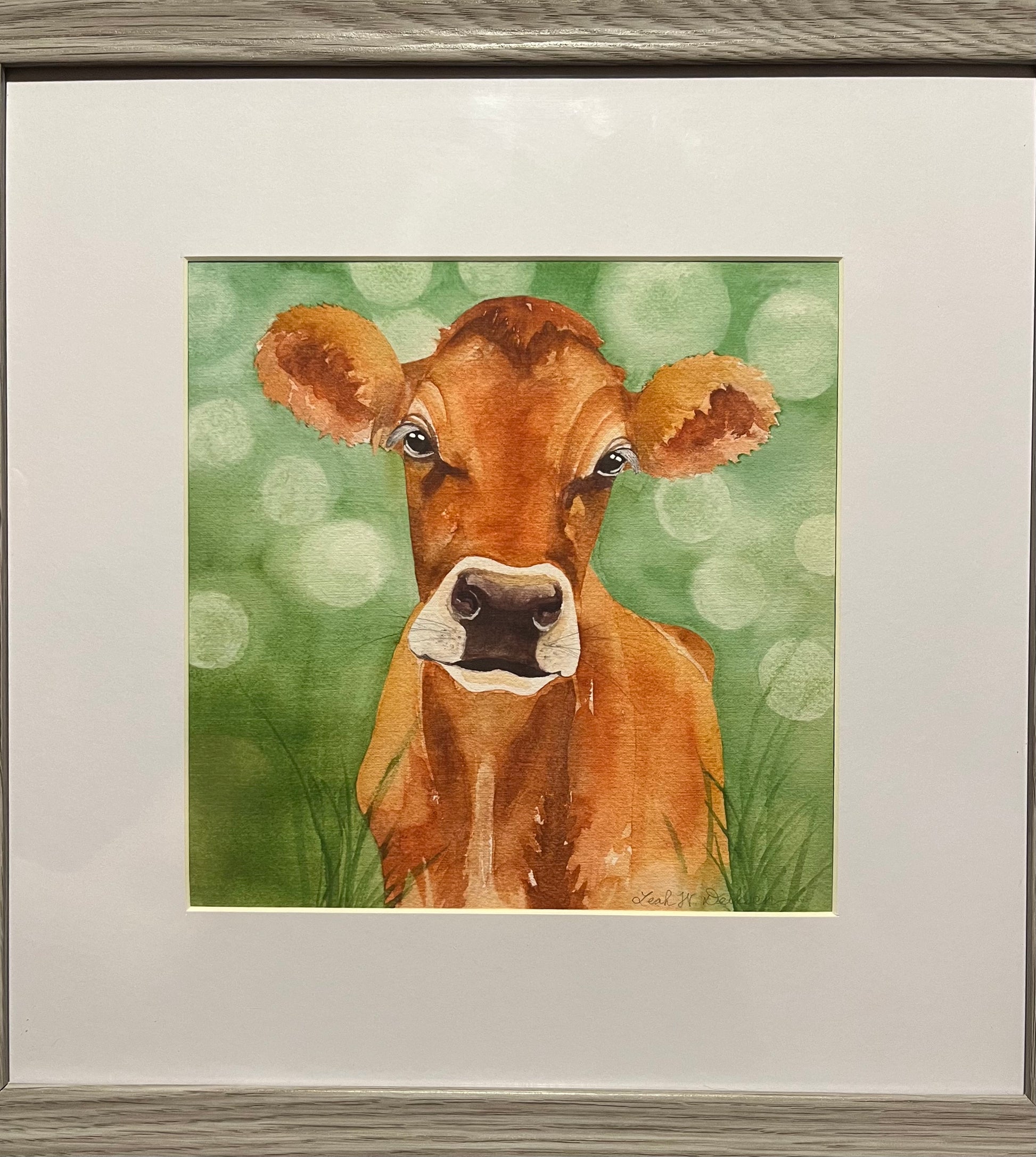 Watercolor painting of a jersey cow with a green painted background in a grey picture frame. 