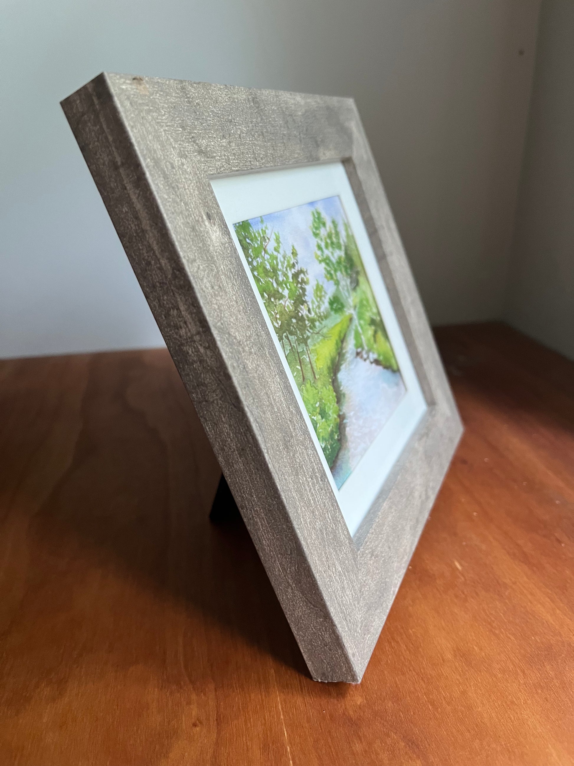 Side view of a grey frame with a watercolor painting of a forest and river