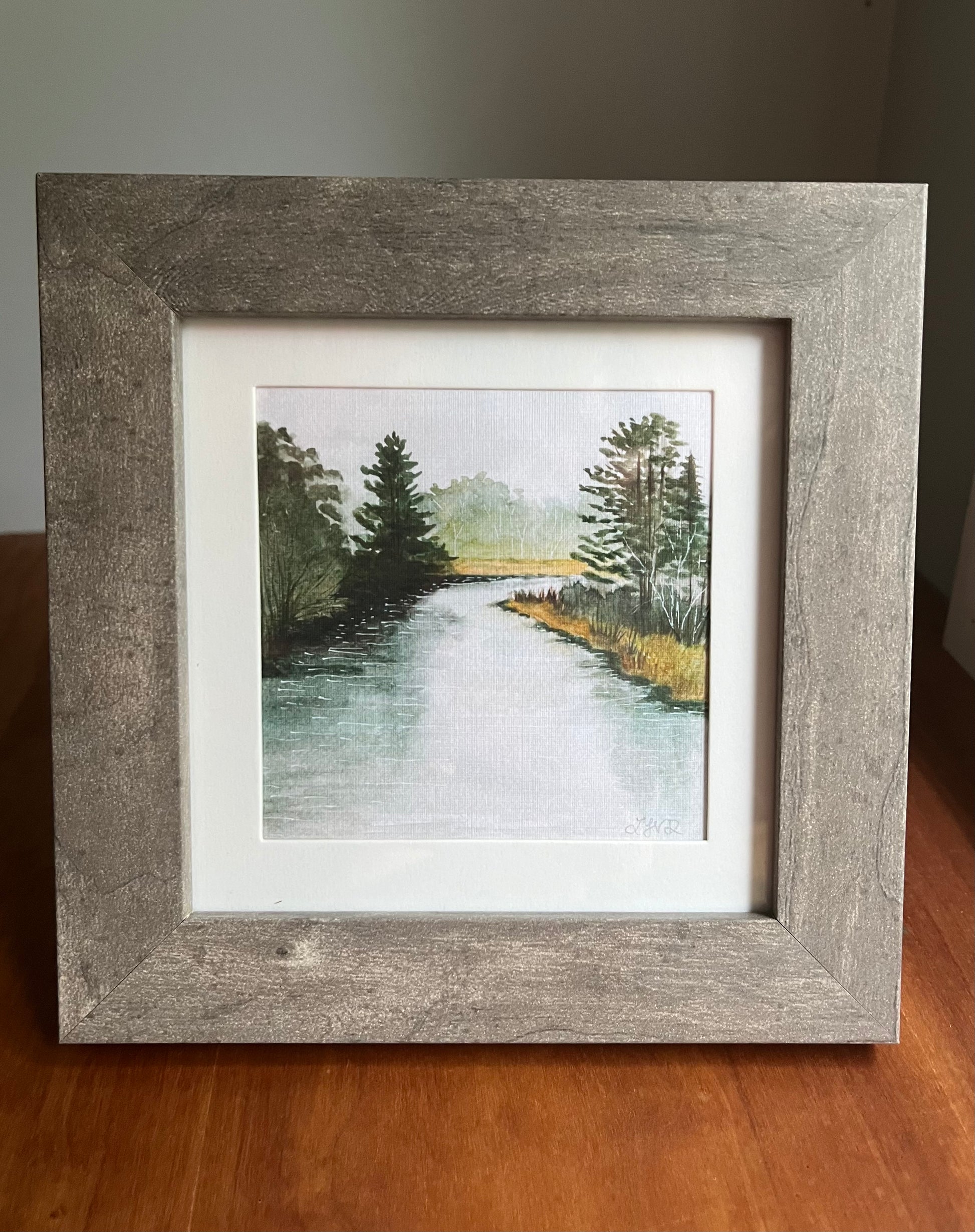Grey picture frame with a white matte holding a watercolor painting of a river and trees