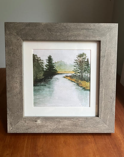 Grey picture frame with a white matte holding a watercolor painting of a river and trees