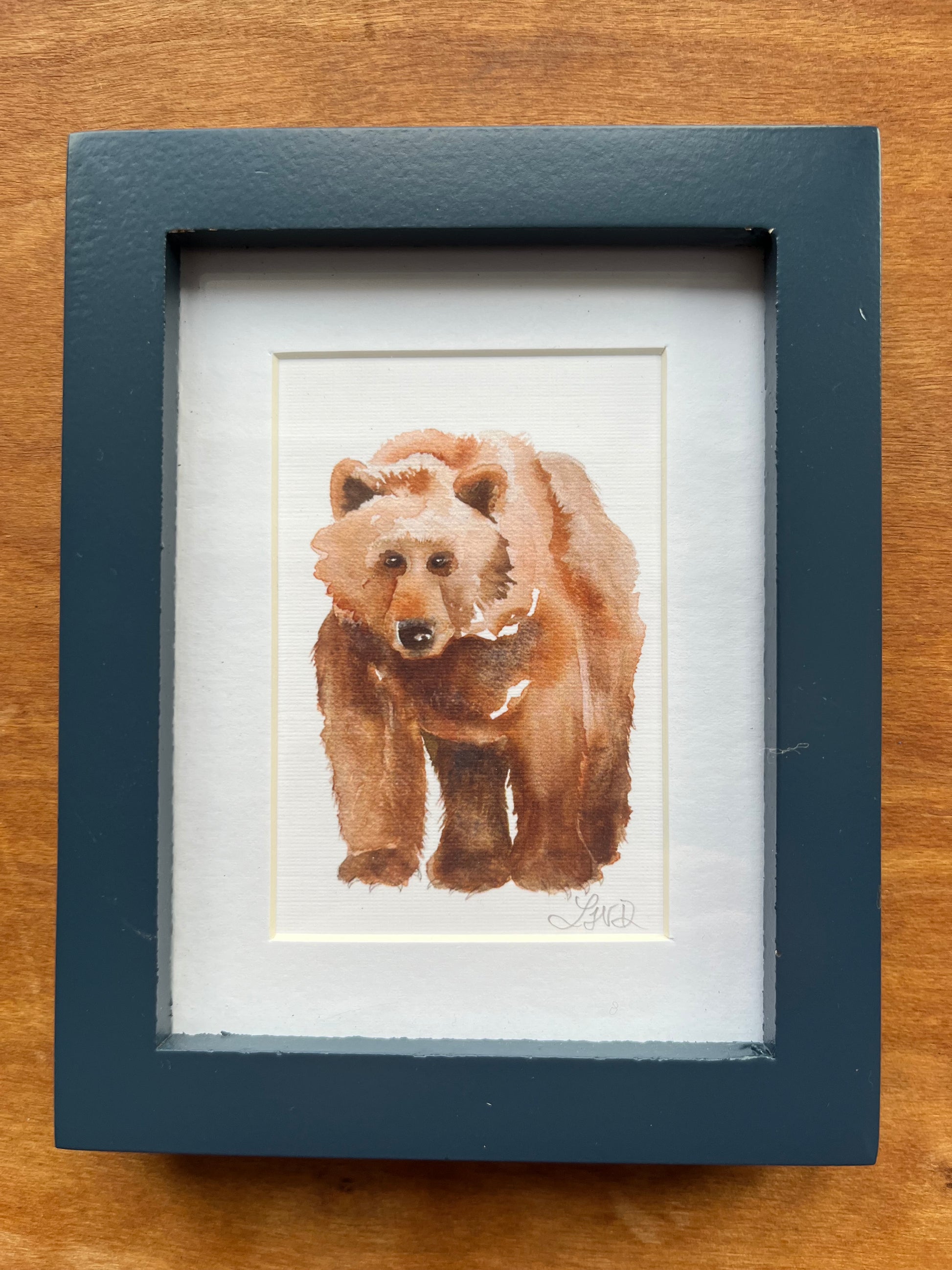 Watercolor painting of a bear in a blue picture frame with a white mat. 