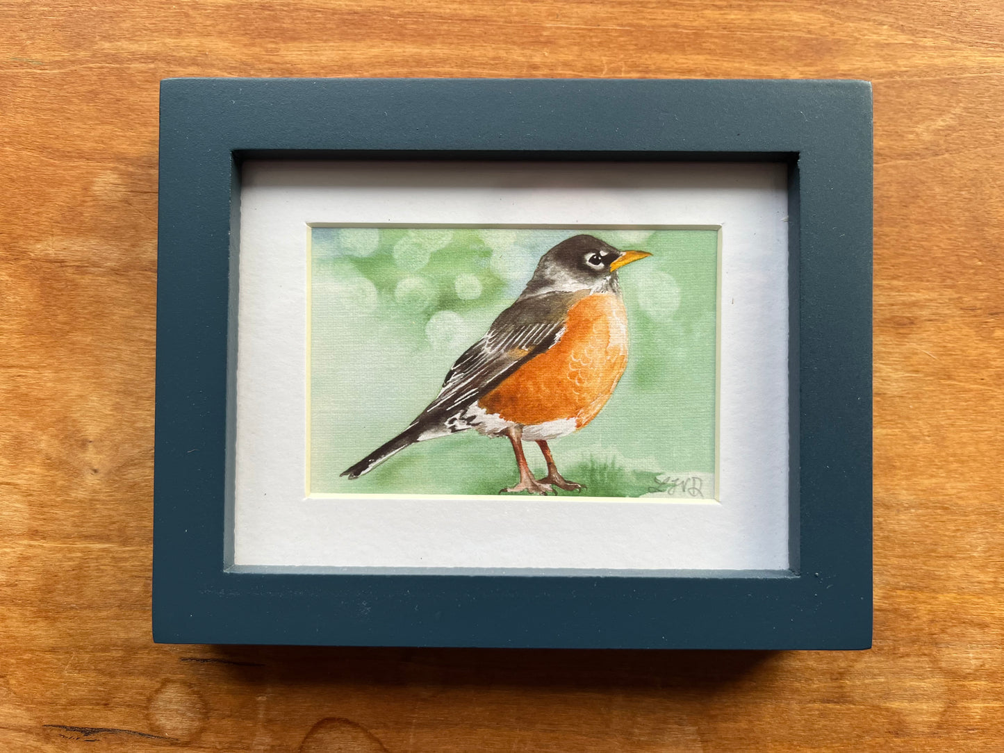 Watercolor painting of a robin with a green background in a blue picture frame. 