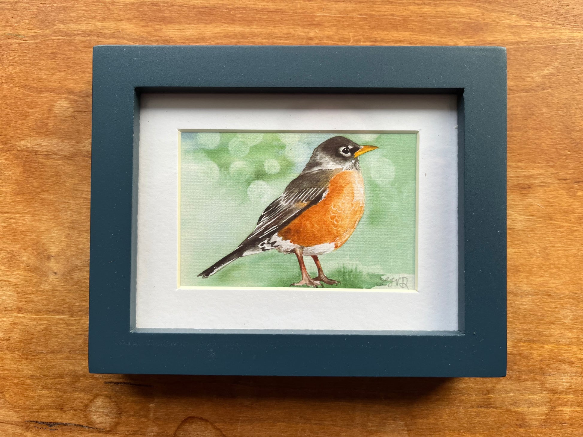 Watercolor painting of a robin with a green background in a blue picture frame. 
