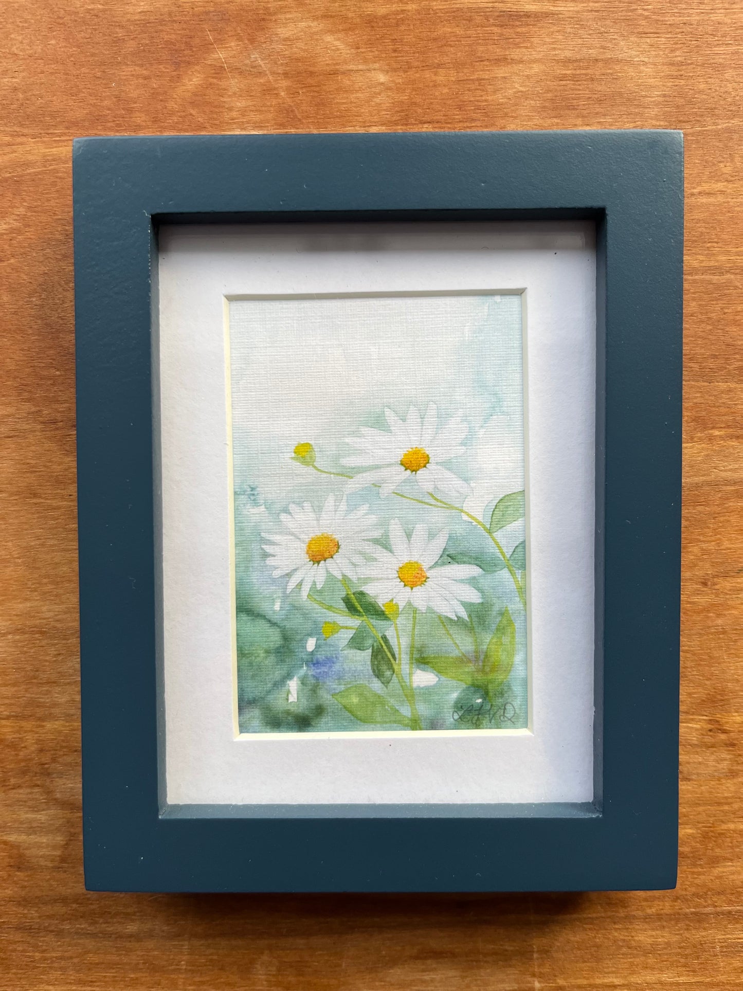 Watercolor painting of daisies in a blue picture frame. 