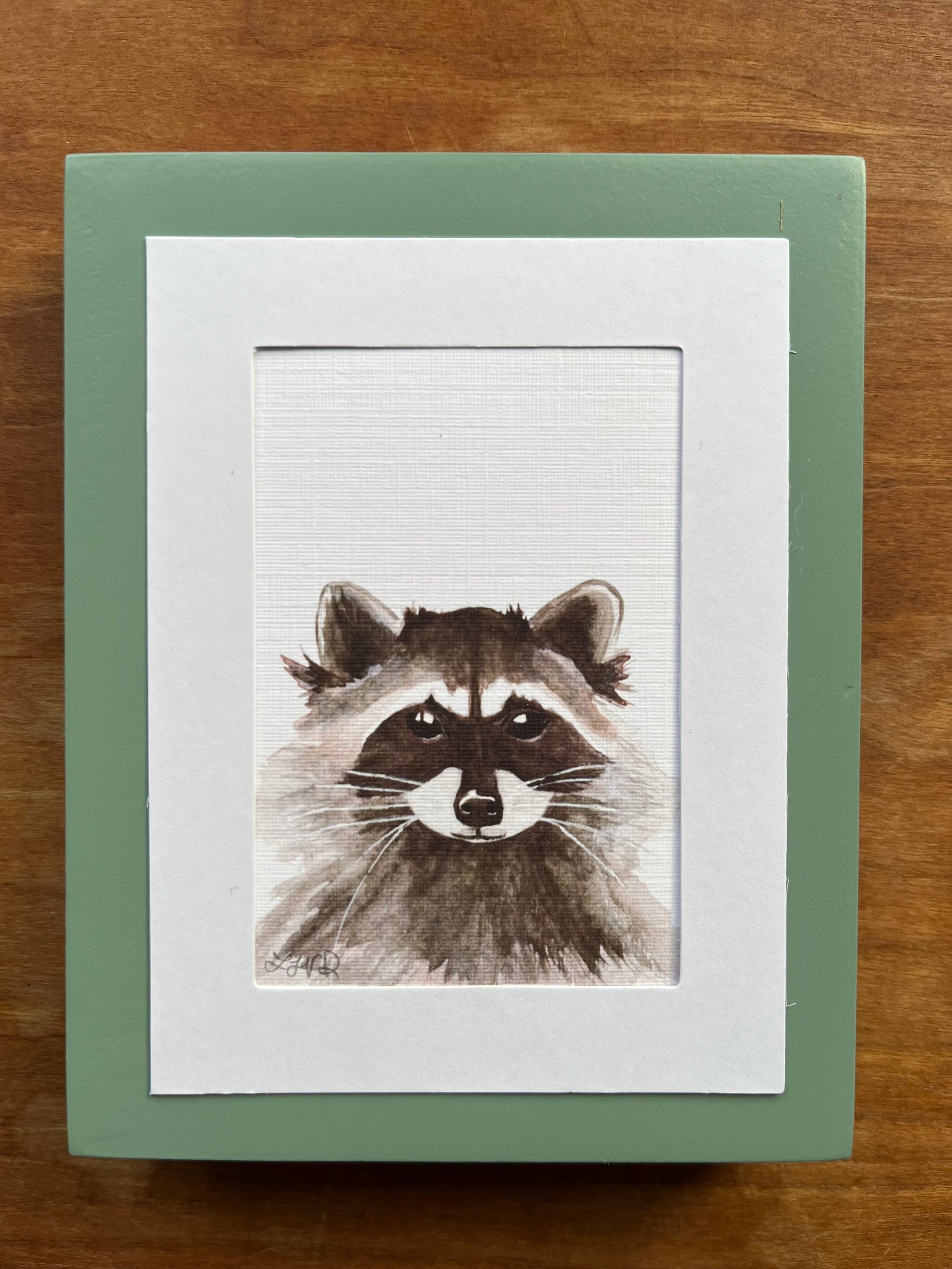 Watercolor painting of a raccoon in a green picture frame. 