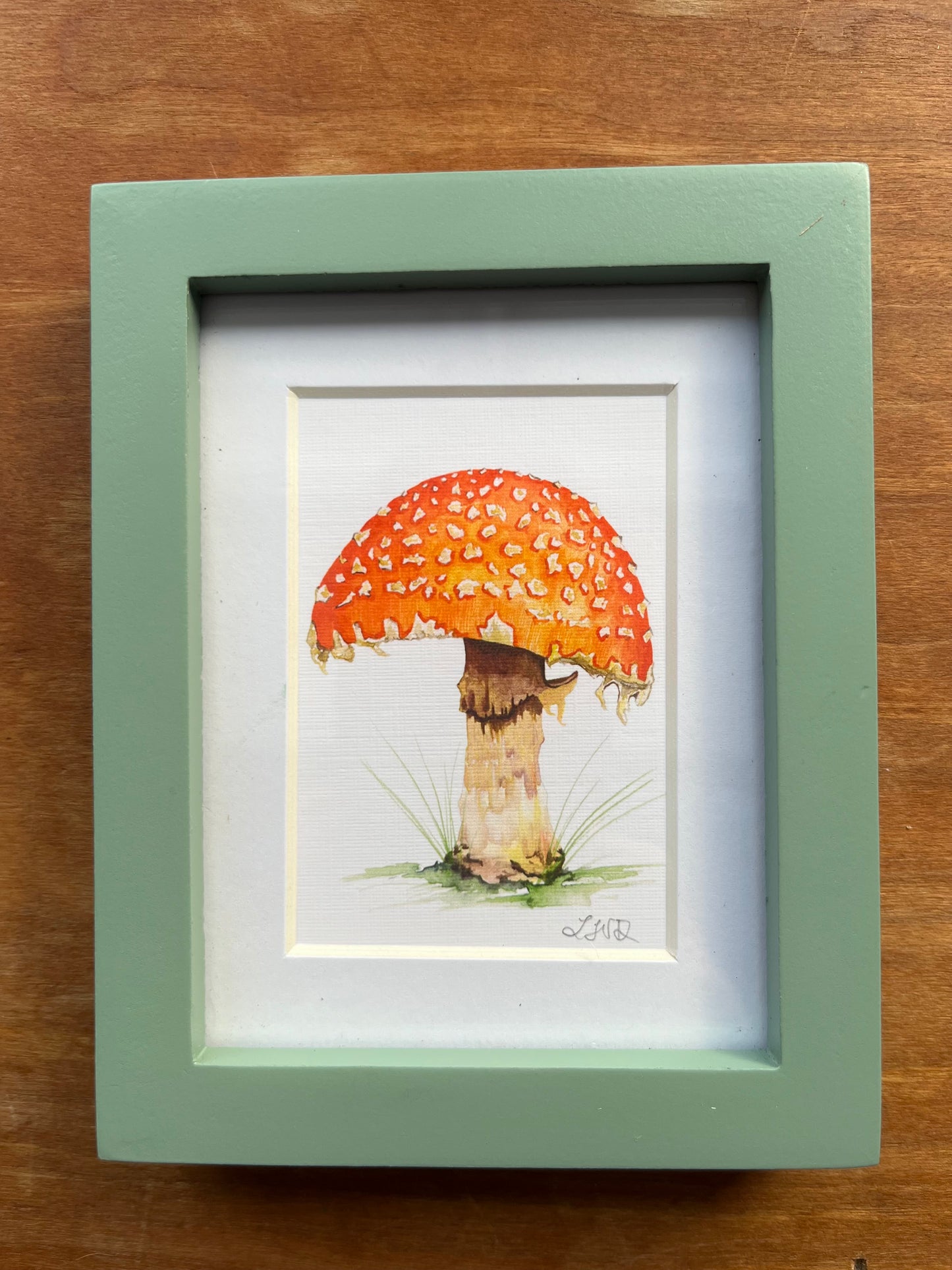 Watercolor fly agaric mushroom painting in a green picture frame. 