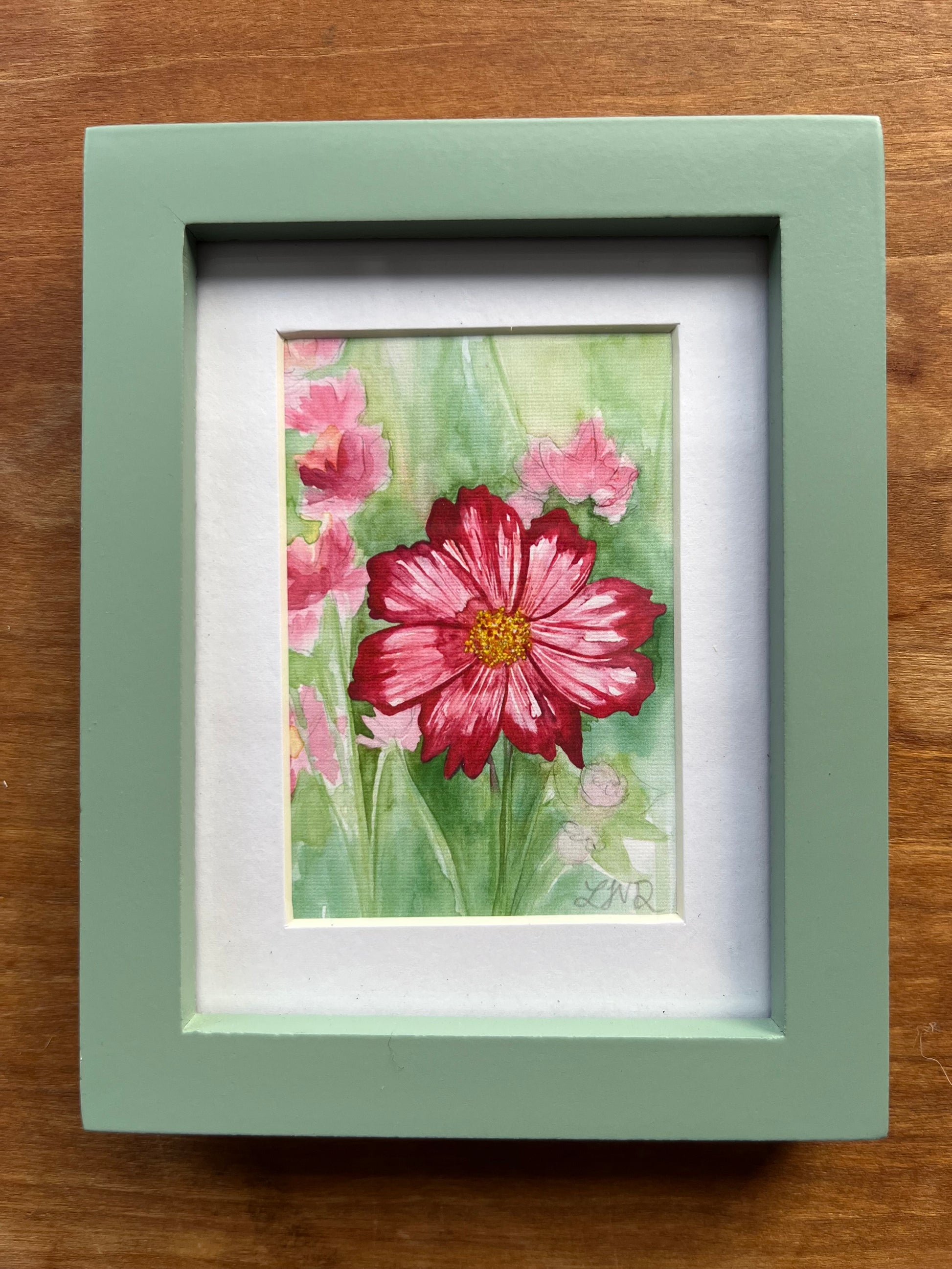 Watercolor painting of a pink cosmos flower in a field in a green picture frame. 