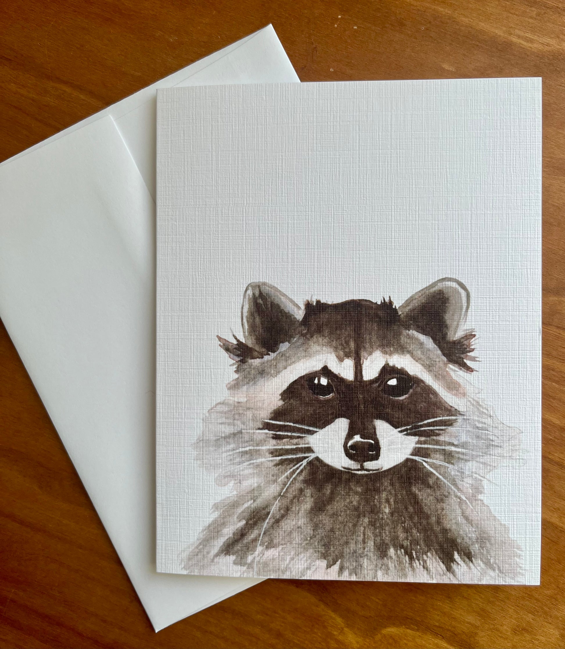 Greeting card with a watercolor painting of a raccoon along with a white envelope.