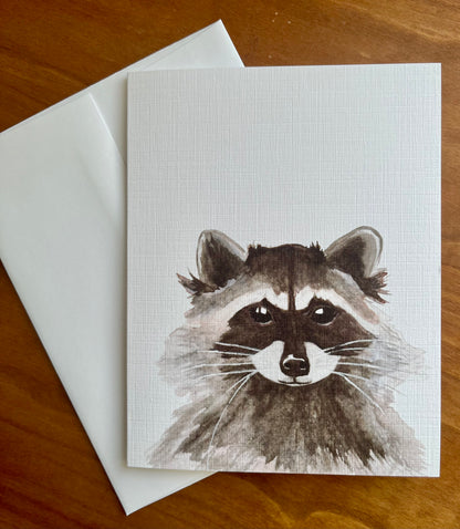 Greeting card with a watercolor painting of a raccoon along with a white envelope.