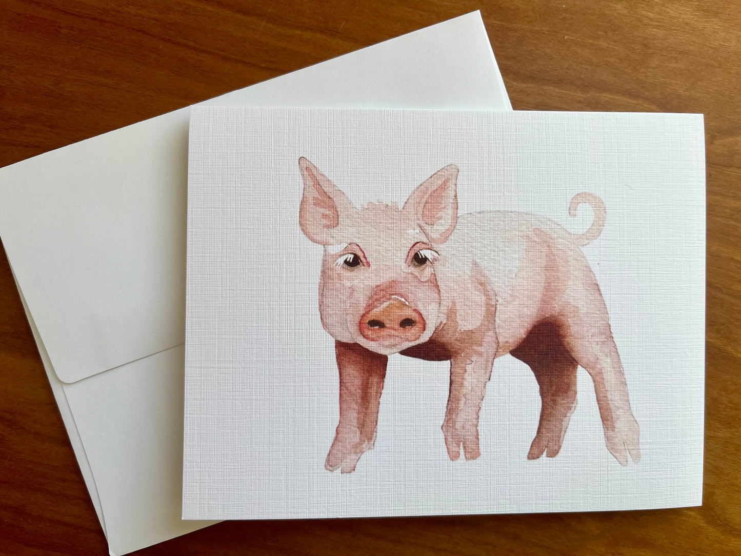 Watercolor painting of a pink pig printed on a greeting card with a white envelope. 