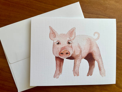 Watercolor painting of a pink pig printed on a greeting card with a white envelope. 
