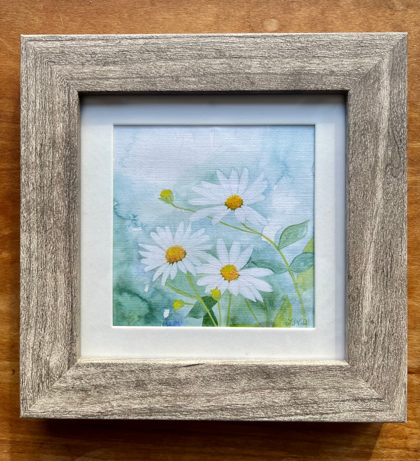 Watercolor painting of daisies in a square grey frame with a white mat. 