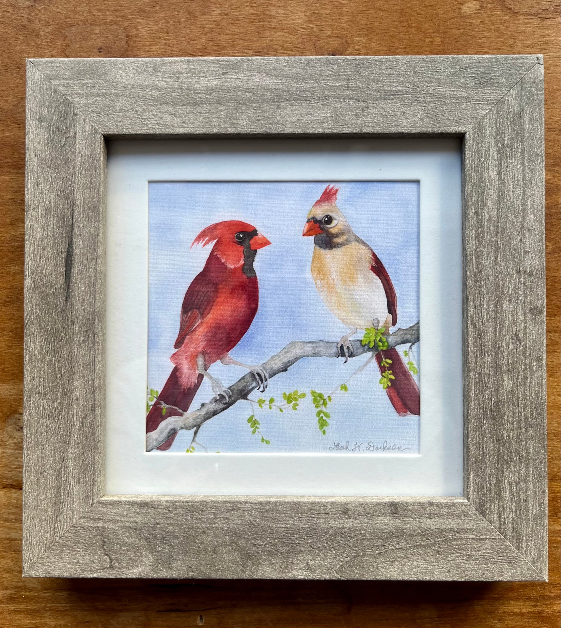 Watercolor painting of a male and female cardinal in a square gray frame with a white mat. 