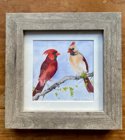 Watercolor painting of a male and female cardinal in a square gray frame with a white mat. 