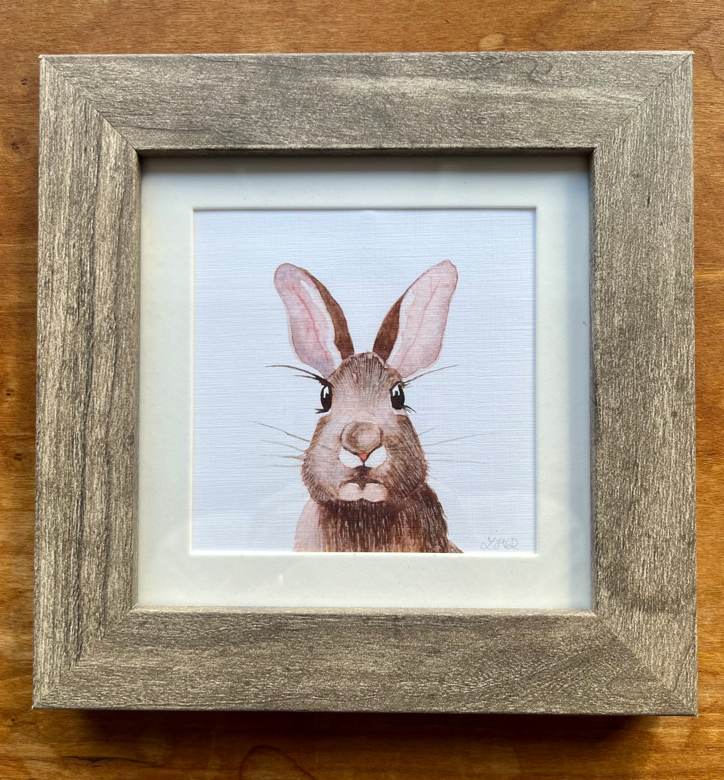 Watercolor painting of a brown rabbit in a square grey frame with a white mat. 