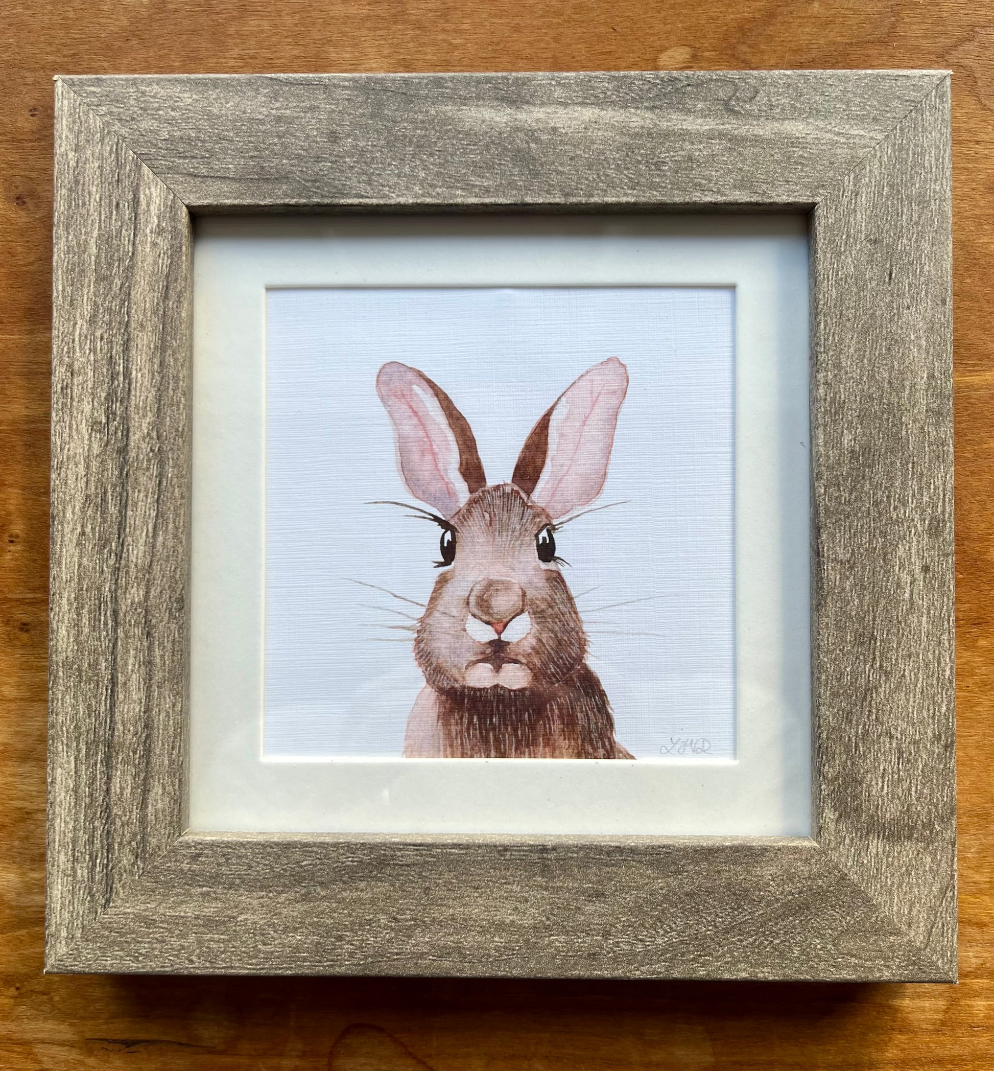Watercolor painting of a brown rabbit in a square grey frame with a white mat. 