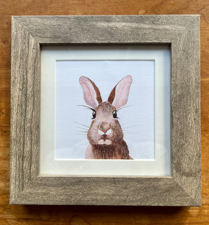Watercolor painting of a brown rabbit in a square grey frame with a white mat. 