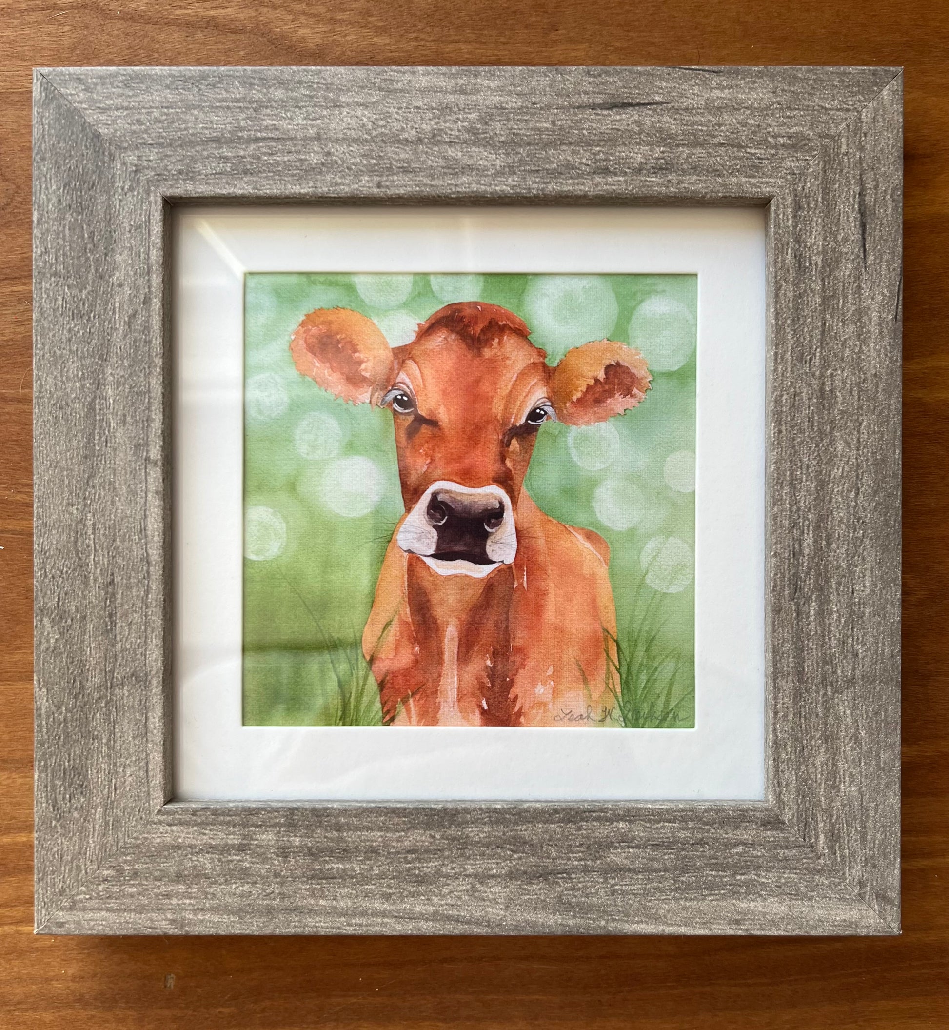 Watercolor painting of a jersey cow in a green field in a square grey frame. 