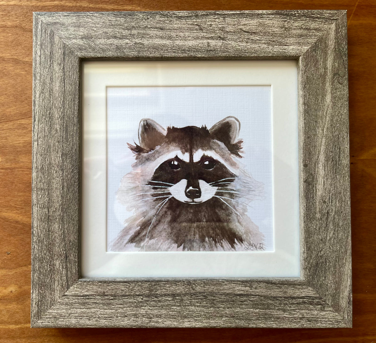 Watercolor painting of a raccoon in a square grey frame. 