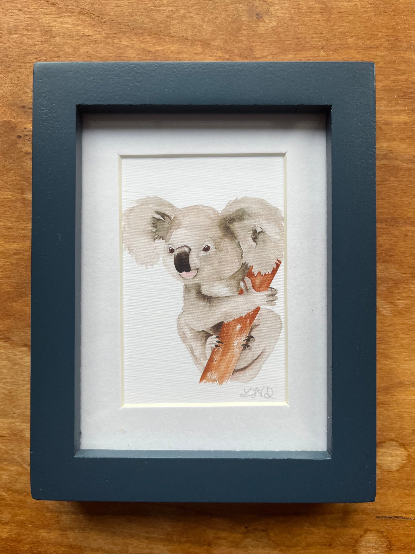 Watercolor painting of a koala holding onto a tree trunk in a blue picture frame. 