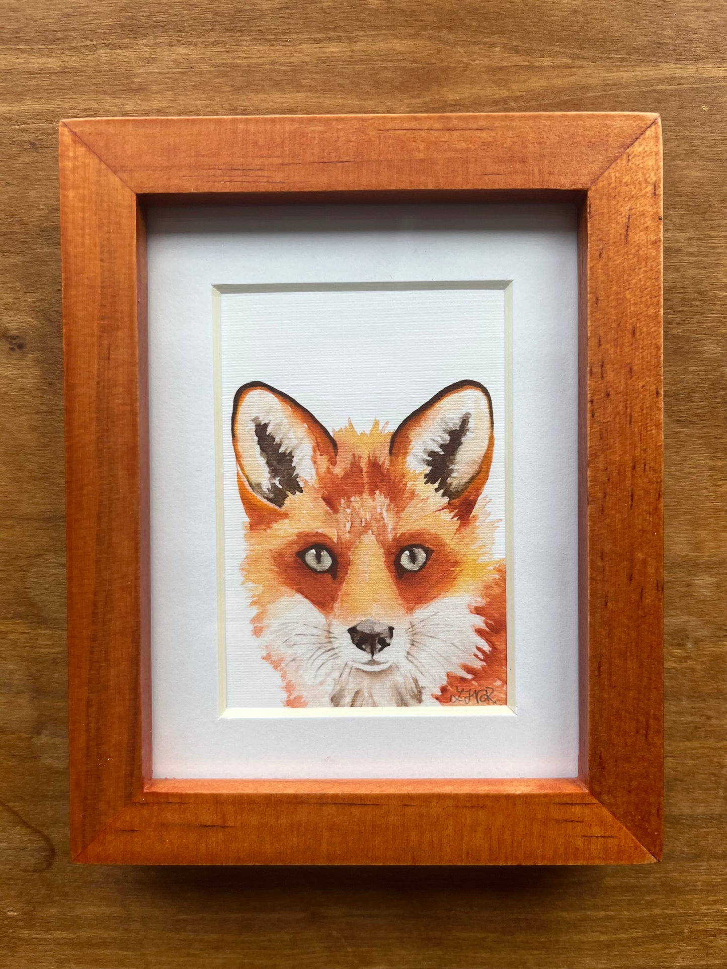 Watercolor painting of a red fox in a brown picture frame. 