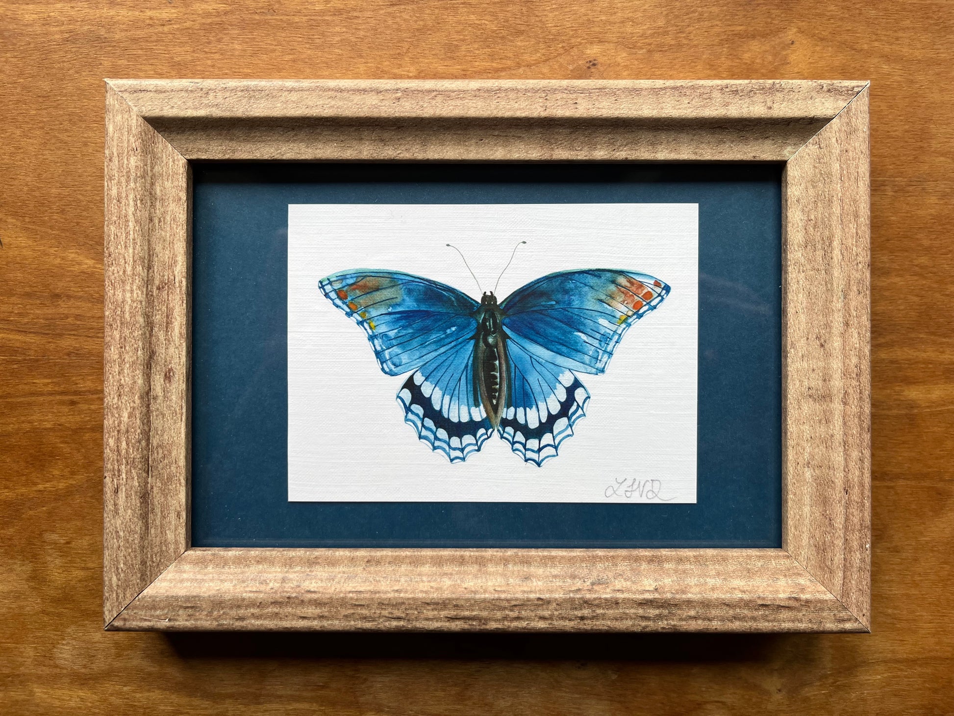 Watercolor painting of a red spotted purple butterfly in a brown frame with a teal blue matte. 