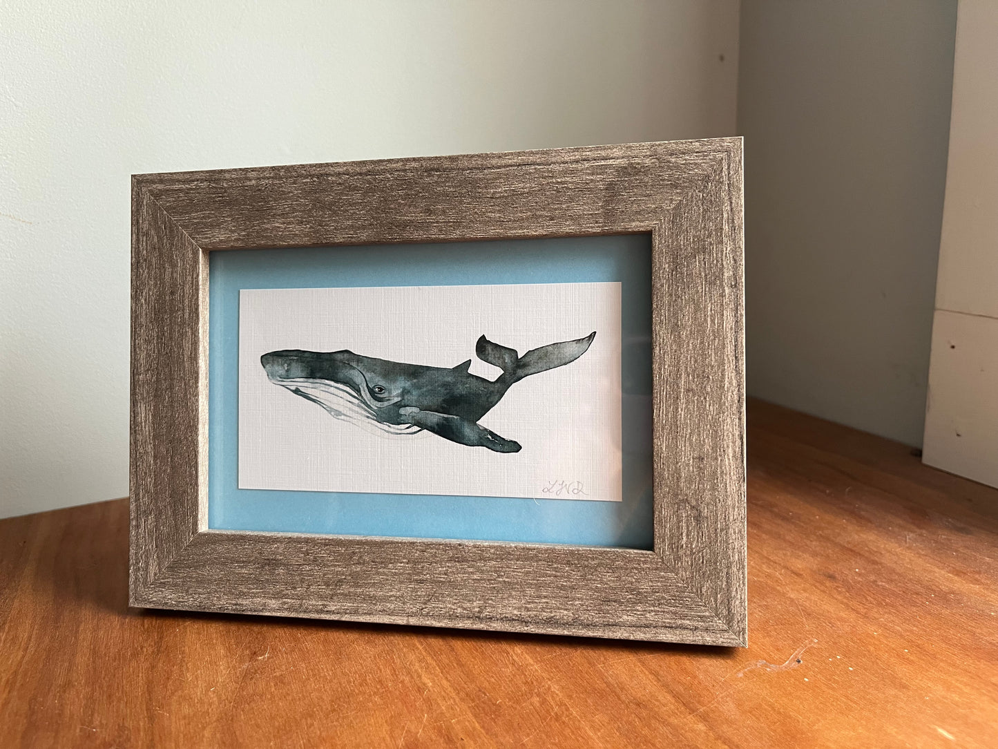 Watercolor whale in a grey wood frame with a light blue matte standing up on a table top. .