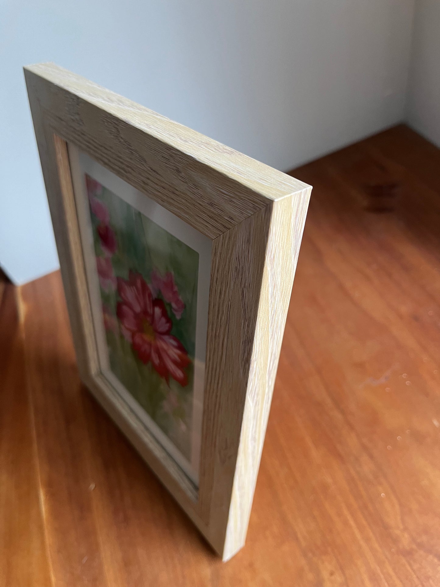 Side view of watercolor painting of pink cosmos flowers in a blonde wood picture frame.