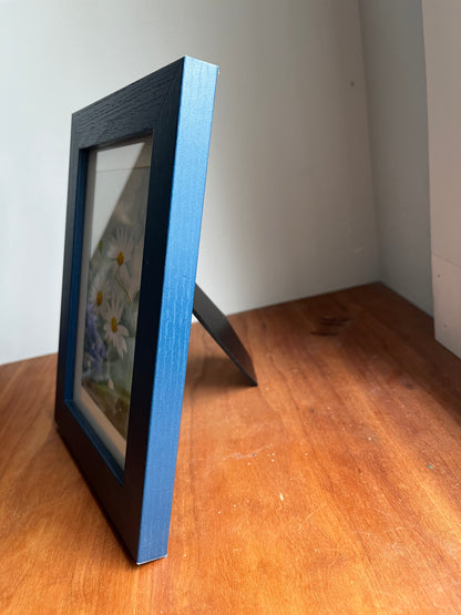 Side view of watercolor daisies in a blue wood picture frame.