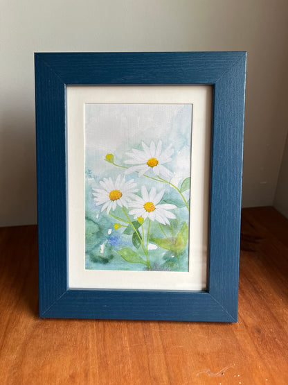 Watercolor daisies in a blue wood picture frame standing up on a table top..