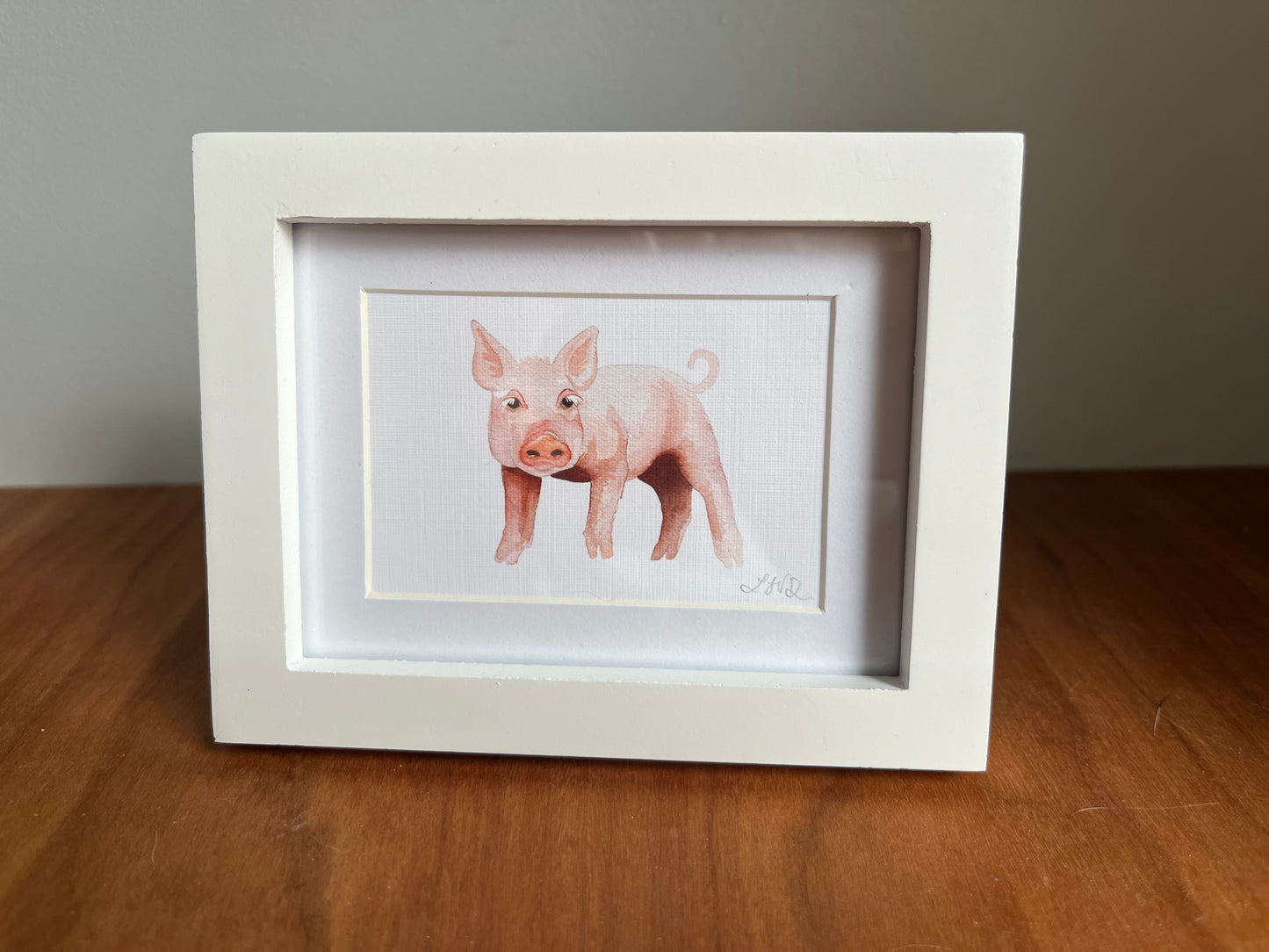 Watercolor pig in a white picture frame with a white matte standing up on a table top.