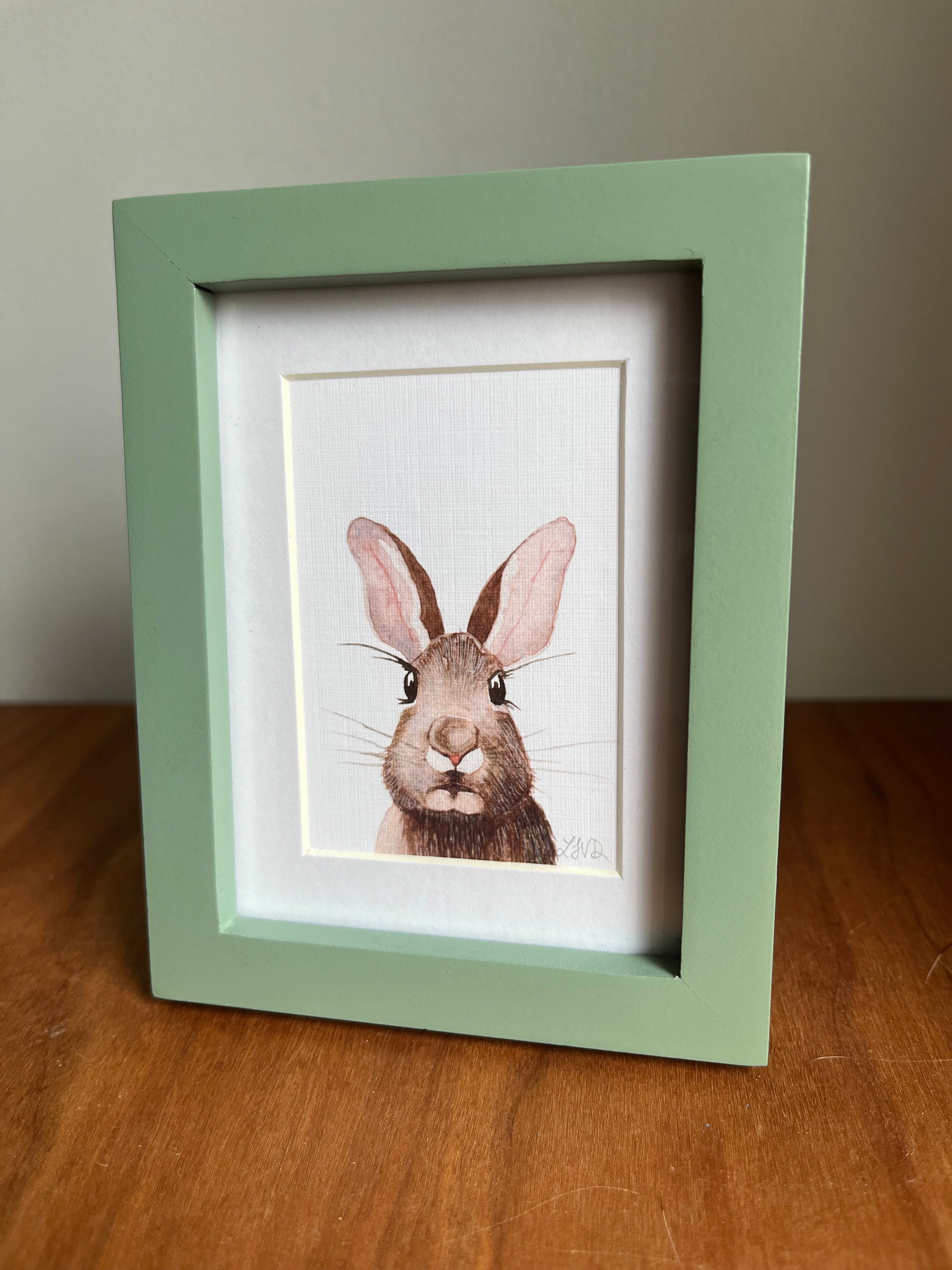 Watercolor rabbit in a green picture frame with a white matte standing up on a table top.