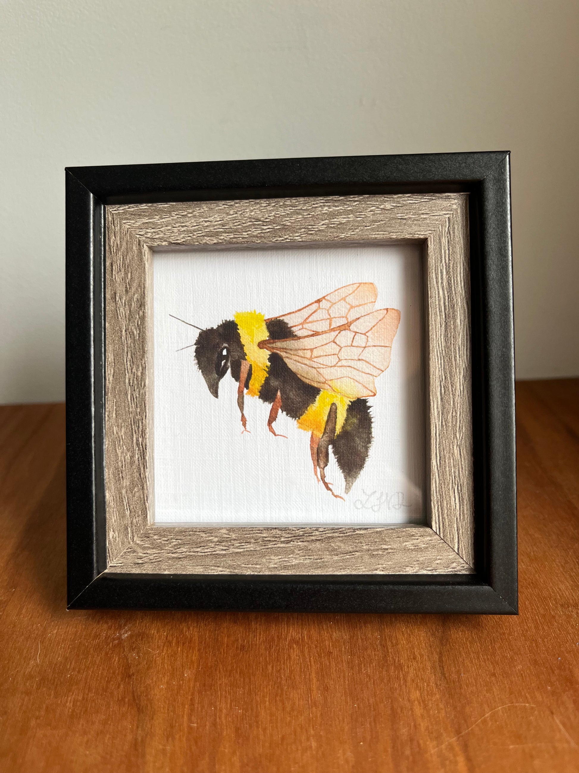 Black and wood square picture frame with a watercolor bee standing on a table top..