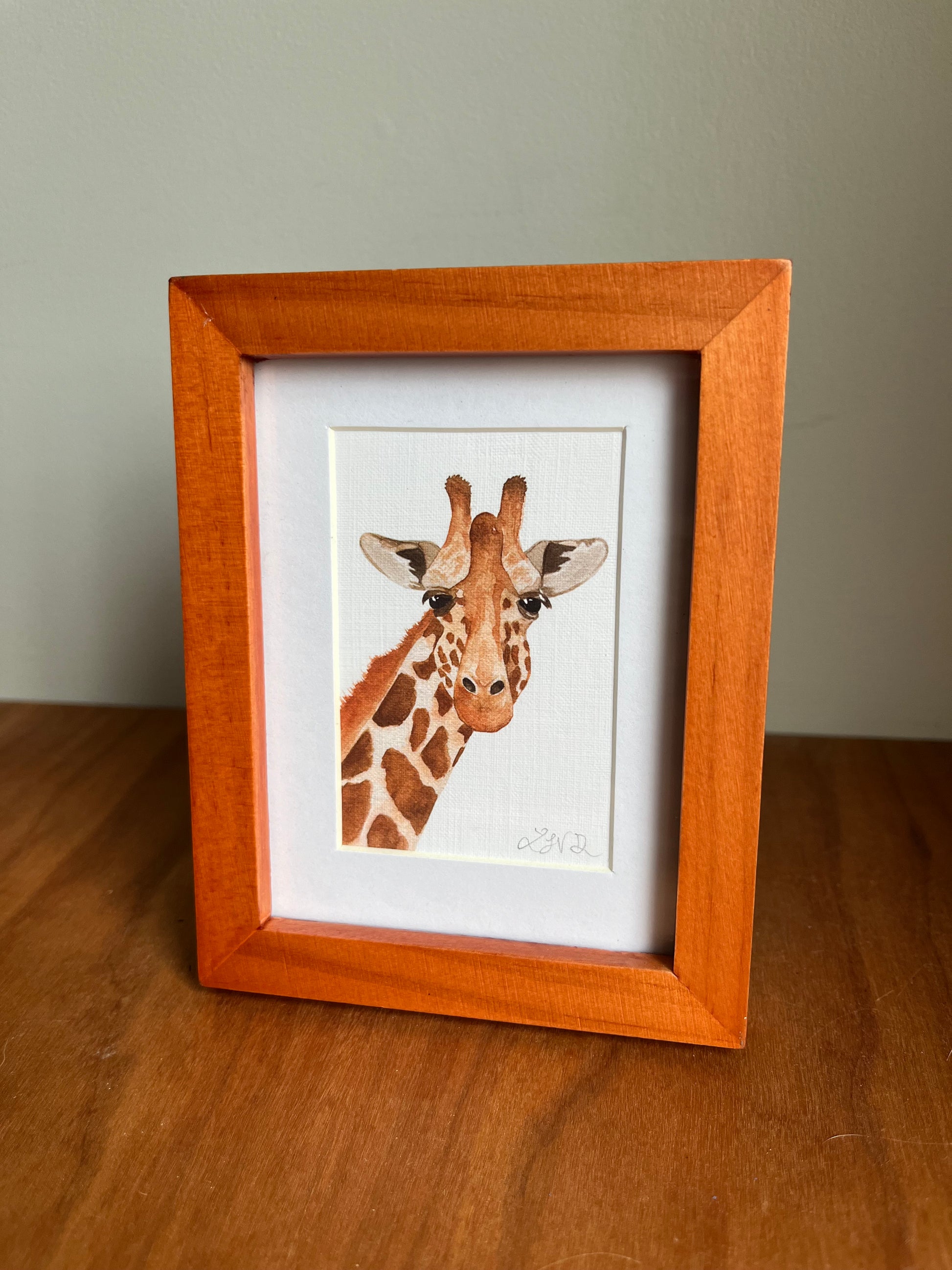 Watercolor giraffe in a honey colored wood picture frame standing up on a table top.